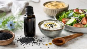 Flašica ulja crnog kima pored crnog semena, jogurta, humusa i sveže salate na kuhinjskoj površini.